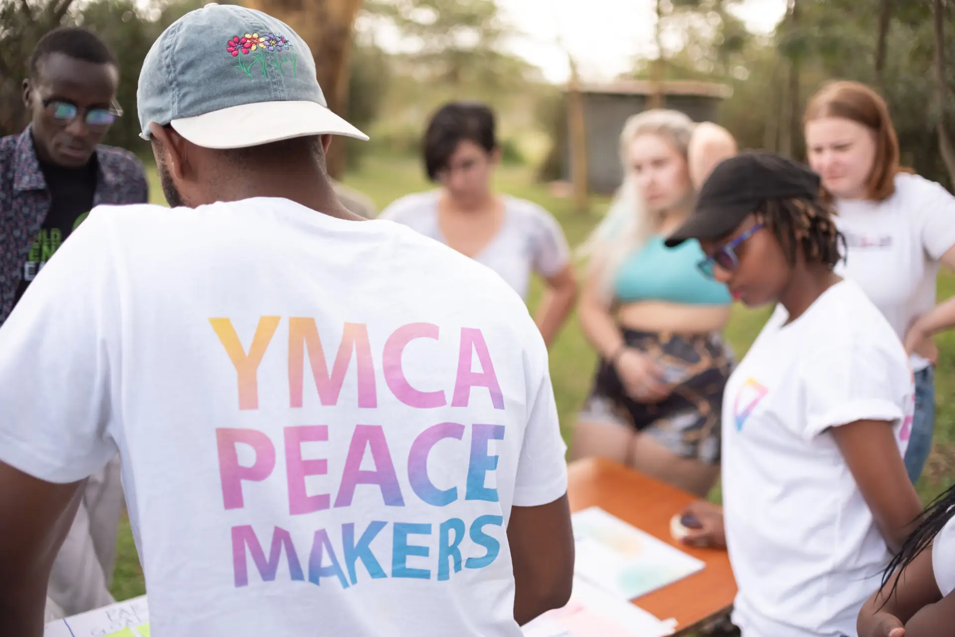 Group of young people outside standing around a table. In the front is a man with his back to the camera in a white t-shirt with the text YMCA PEACEMAKERS on the back in a pastel gradient